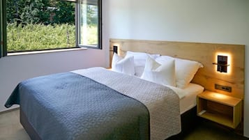 Un dormitorio con cama, mesita de noche, luz adosada a la pared y ventana con vistas a zonas verdes.