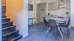 A modern kitchen with a dining area, featuring a staircase, grey tiled flooring, and minimalist cabinetry.