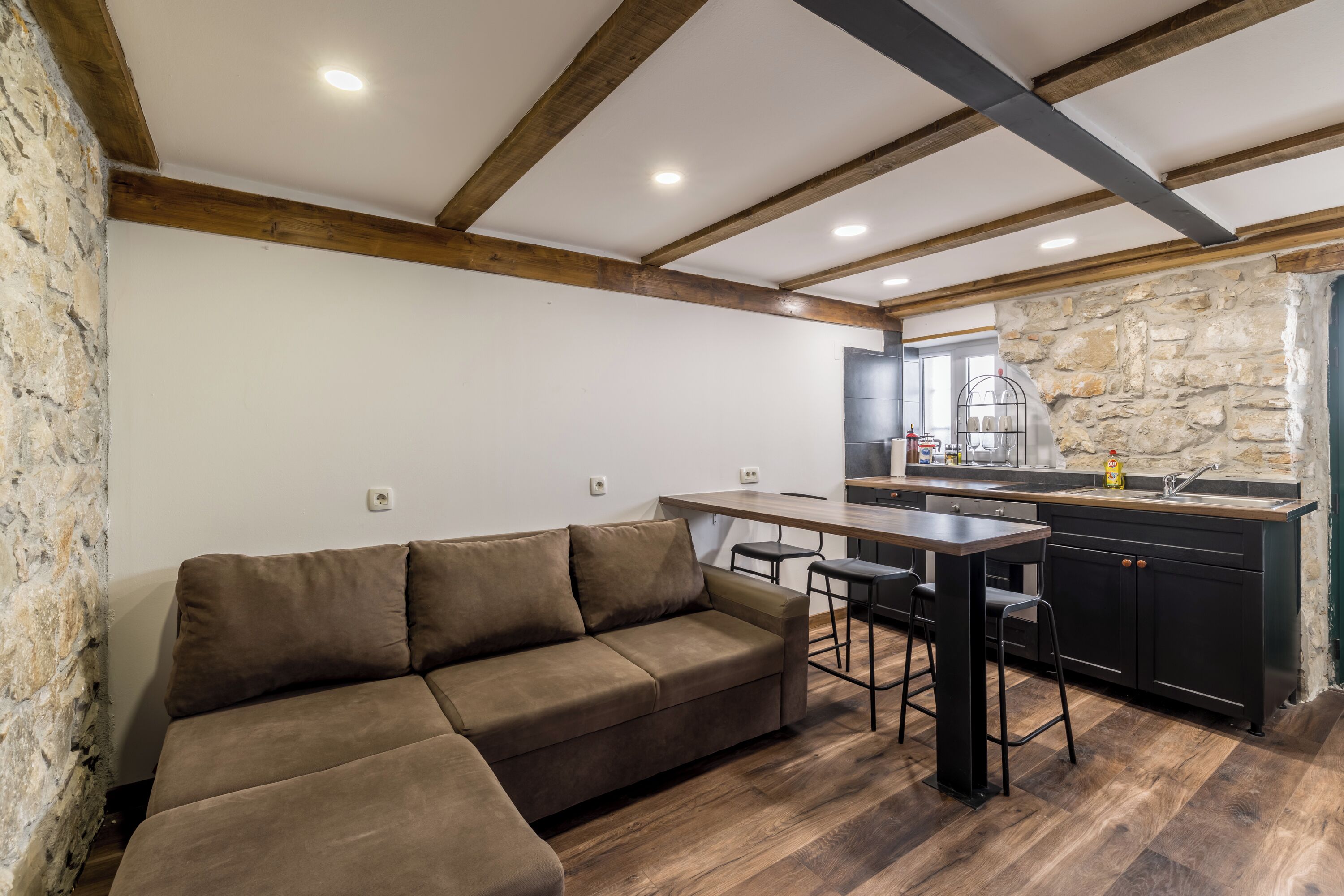 A modern kitchen with a stone accent wall, a wooden ceiling, and a brown sofa in a living area.