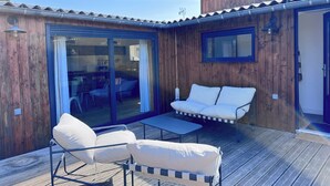 Een houten terras met een schuifdeur naar een eethoek, een zithoek met een tafel, en een klein raam.