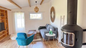 Een woonkamer met een bank, een blauwe fauteuil, een houten boekenkast, een open haard en een raam met uitzicht op bomen.