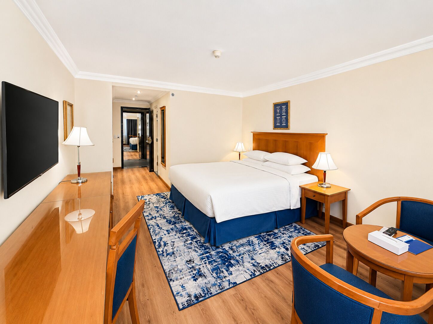 A hotel room with a bed, bedside tables, a desk, a chair, a TV, and a patterned rug.