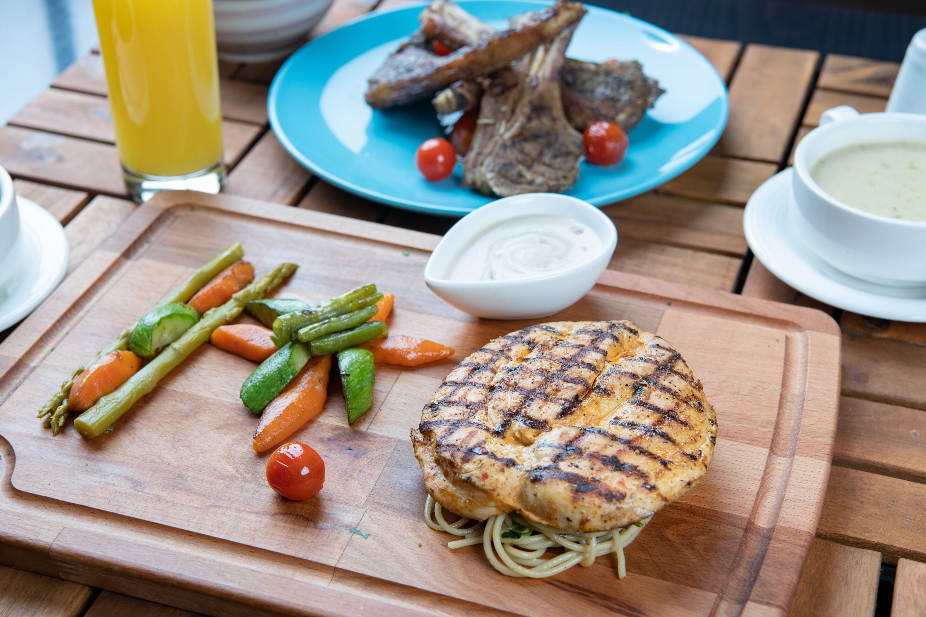 A wooden table with a plate of food, including grilled chicken, vegetables, and a bowl of sauce.