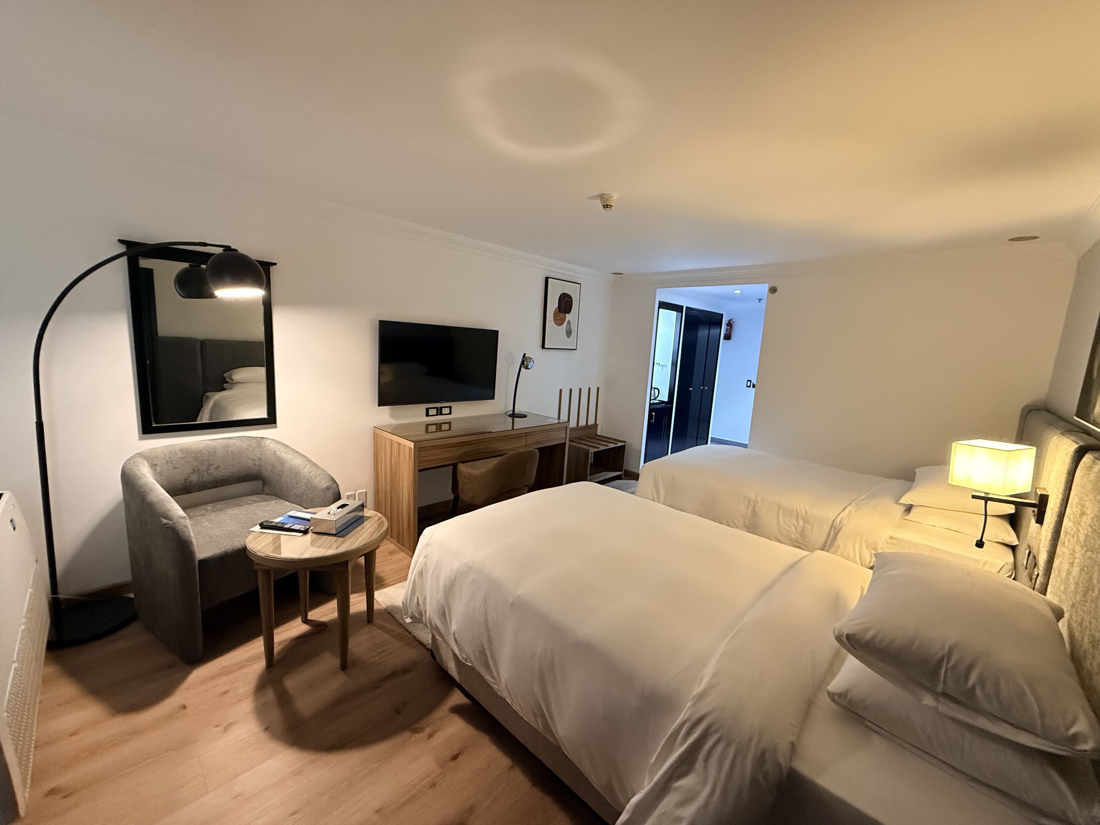 A hotel room with a bed, a desk with a TV, a chair, a lamp, and a door leading to a bathroom.