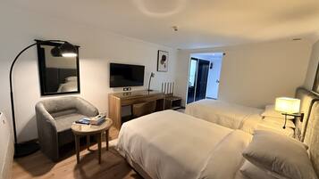 A hotel room with a bed, a desk with a TV, a chair, a lamp, and a door leading to a bathroom.