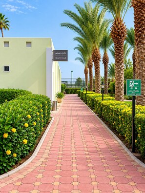 Un chemin bordé de palmiers et de fleurs jaunes, qui mène à un bâtiment portant l’inscription « Club de santé ».