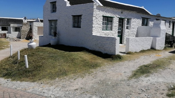 A white building with a sloped roof, a small porch, and a stone wall. - Quaint 1 Bedroom Apartment With sea View (Arniston)