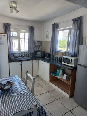 A compact kitchen with a microwave, toaster, and refrigerator. There is a window with curtains, a tiled backsplash, and a table with a checkered tablecloth. - Quaint 1 Bedroom Apartment With sea View (Arniston)