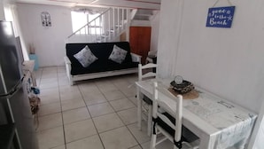 A room with a sofa, a dining table with chairs, a refrigerator, and a staircase. - Quaint 1 Bedroom Apartment With sea View (Arniston)