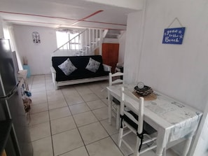 A room with a sofa, a dining table with chairs, a refrigerator, and a staircase.