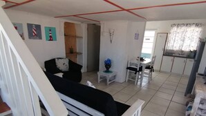 A living room with a sofa, a dining table with chairs, a kitchen area, and a staircase. - Quaint 1 Bedroom Apartment With sea View (Arniston)