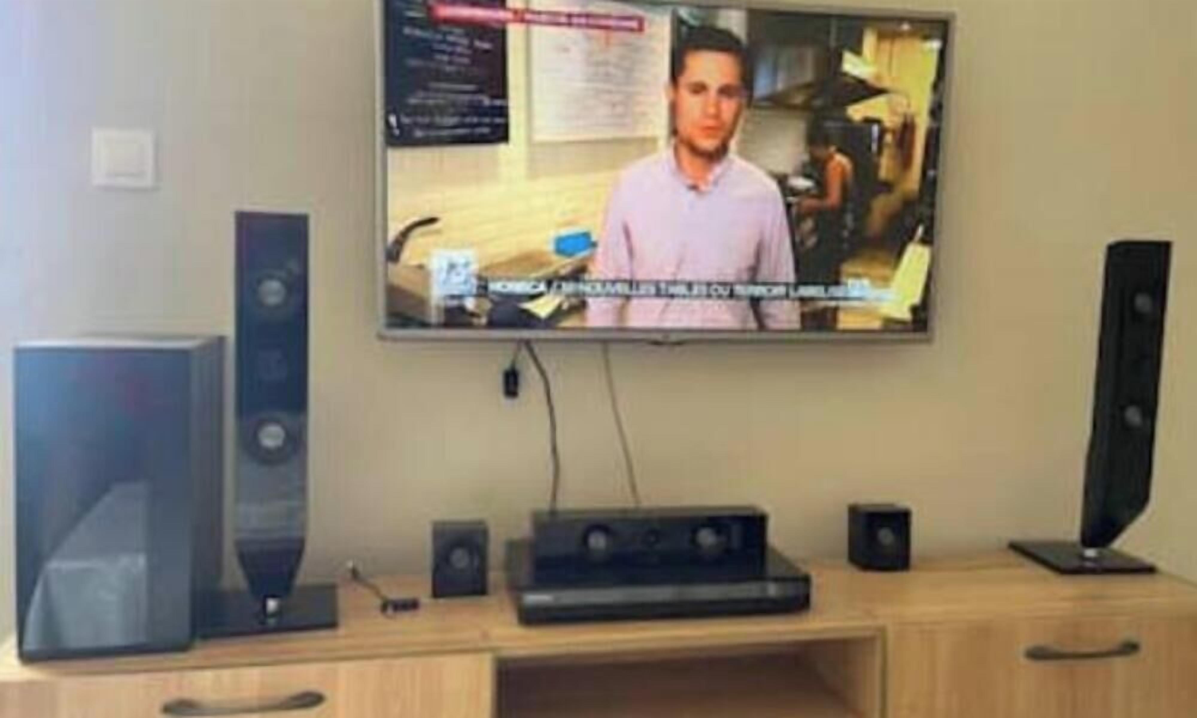 Uma televisão na parede, uma secretária de madeira com um computador e dois alto-falantes.