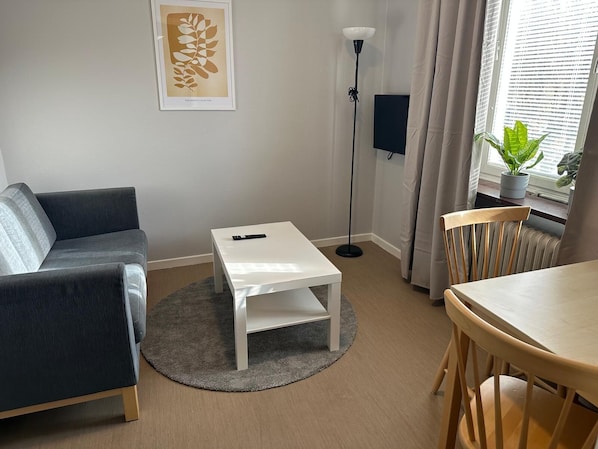 Ett modernt vardagsrum med en grå soffa, ett vitt soffbord, en golvlampa, ett matbord med stolar och ett fönster med gardiner.
