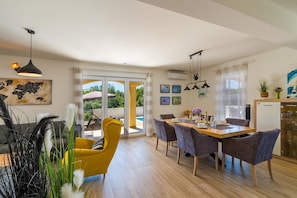 A dining area with a wooden table and chairs, a yellow armchair, and a view of the outdoors through glass doors. - Villa Florami – Heated Pool, Sea Views and Peaceful Location (Krnica)
