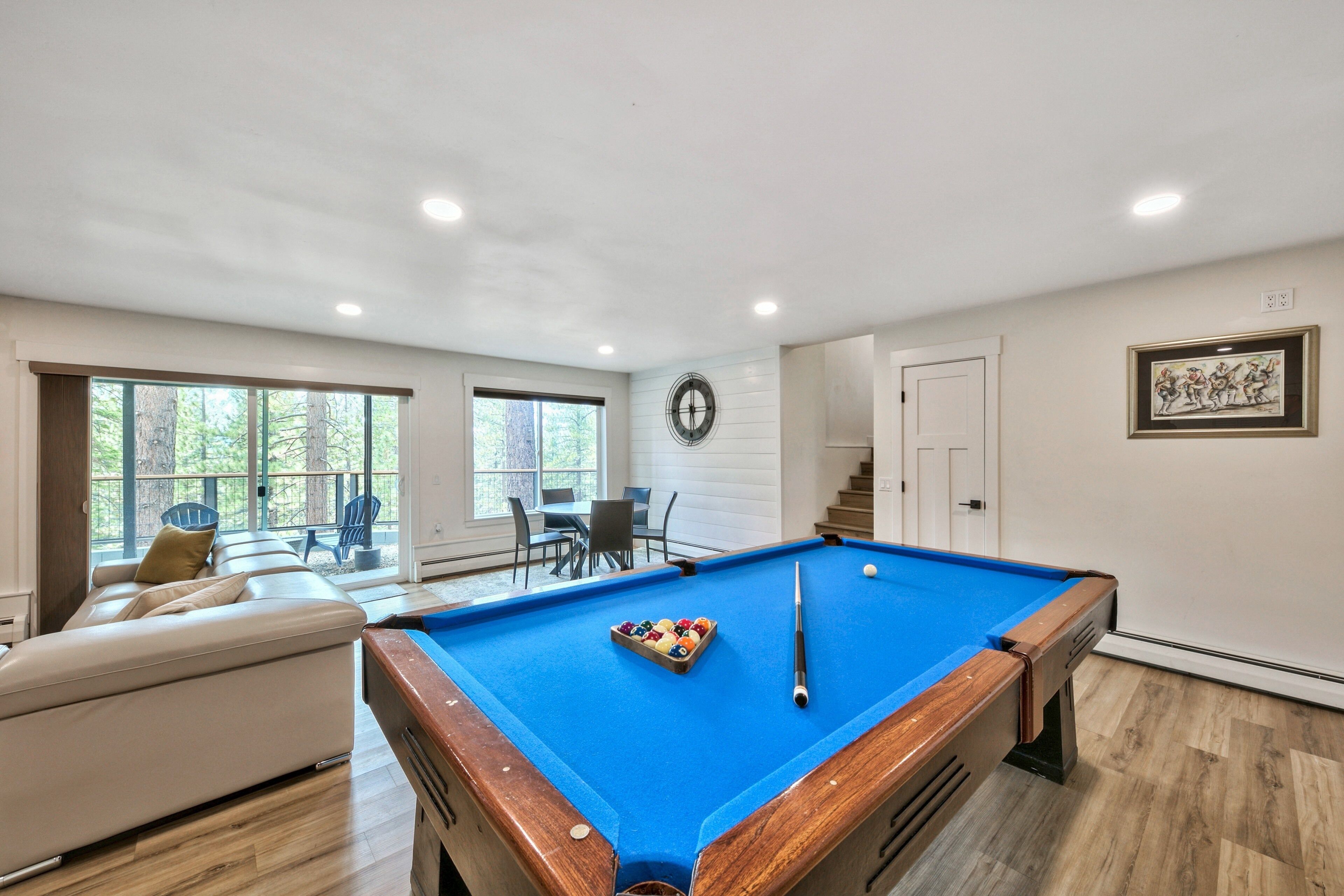 A pool table with a blue felt surface, a pool cue, and a set of balls. There is a white sofa with a cushion, a dining table with chairs, and a window with a view of trees.