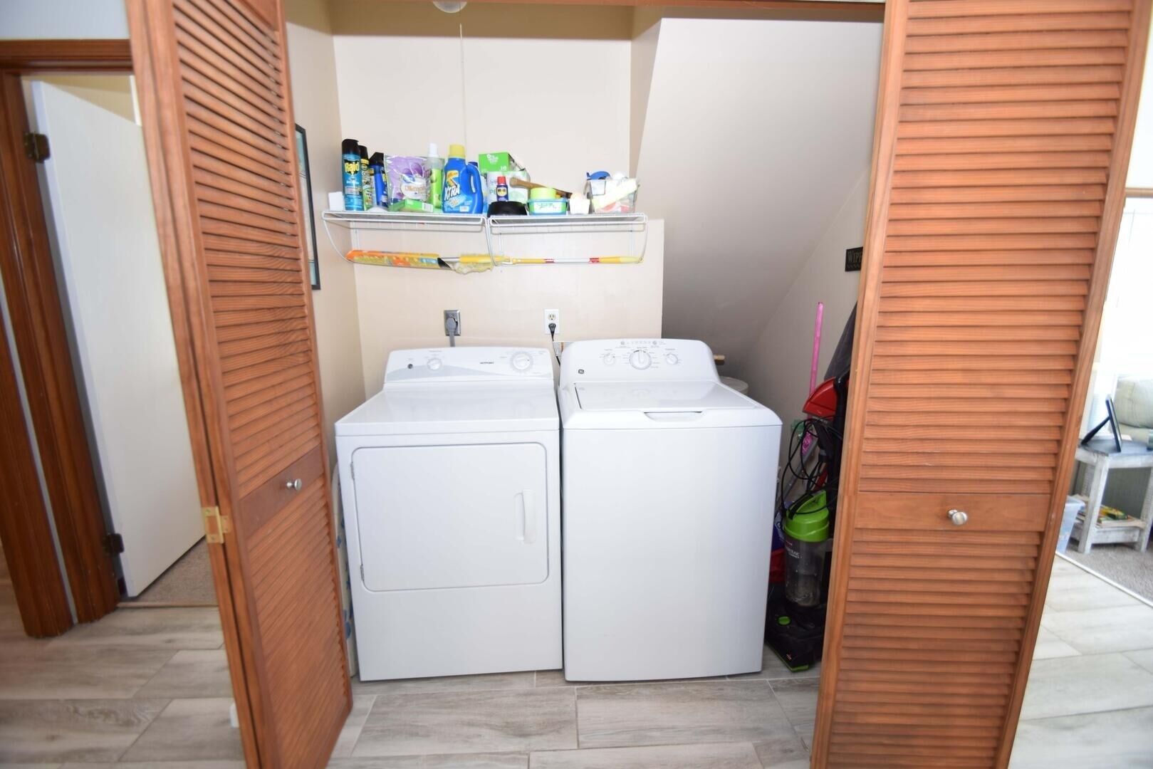 A laundry room with two white washing machines, a shelf with cleaning supplies, and a door leading to another room.