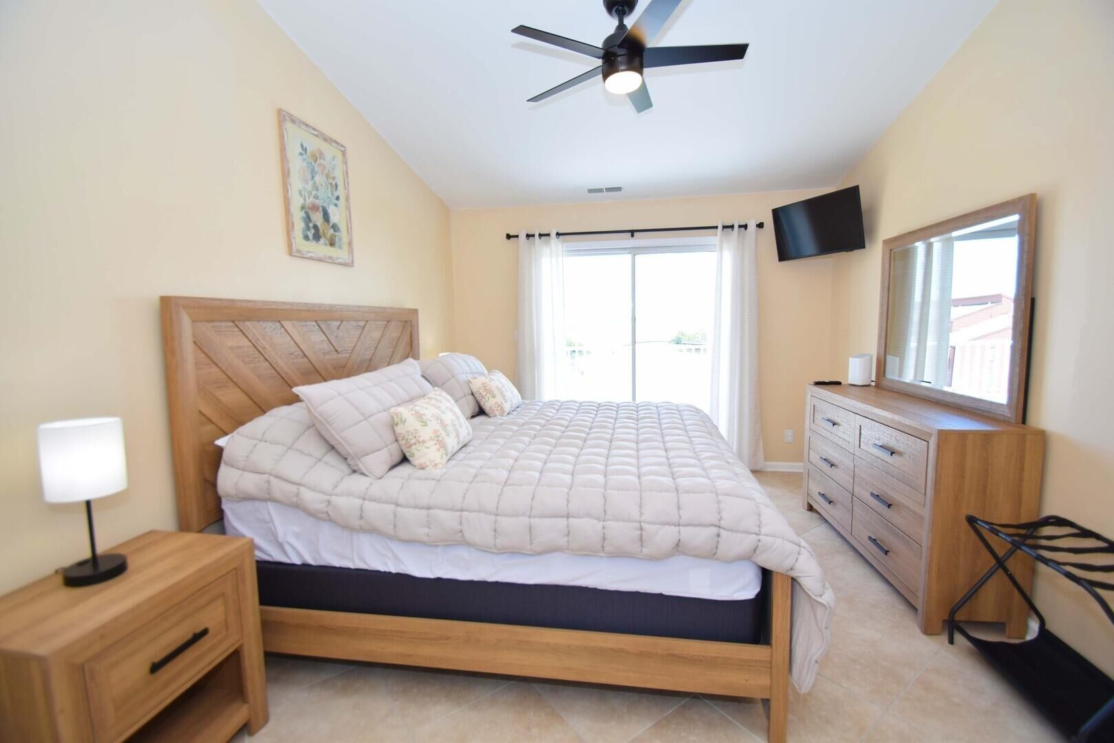 A bedroom with a bed, a nightstand, a dresser, a ceiling fan, a TV, and a window with curtains.