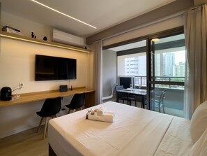 A hotel room with a bed, a desk with a TV, a chair, a table with a glass, and a balcony with a view of buildings. - 28m² 2 People Pool Gym (São Paulo)