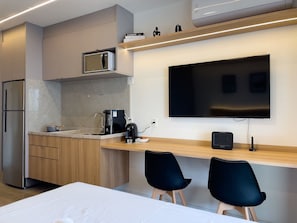 A modern kitchen with a built-in microwave, a coffee maker, and a flat-screen TV mounted on the wall. There are two black chairs and a wooden table. - 28m² 2 People Pool Gym (São Paulo)