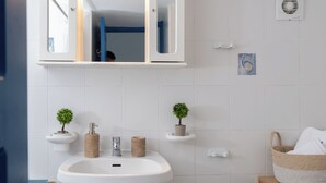 Ein Badezimmer mit einem weißen Waschbecken, zwei kleinen Topfpflanzen, einem weißen Handtuch und einem Korbwannen-Korb.