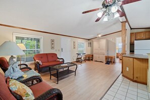 A spacious living area with a red sofa, wicker furniture, a ceiling fan, and a kitchen area with wooden cabinets.