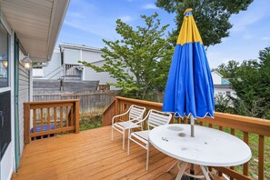 Eine Holzterrasse mit einem runden Tisch, zwei Stühlen und einem Sonnenschirm, im Hintergrund ein Haus zu sehen.