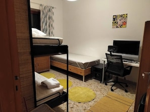 A dormitory room with bunk beds, a desk with a computer, and a chair.