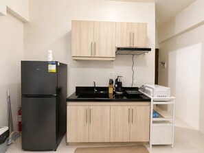 Een kleine keuken met een zwarte koelkast, houten kastjes, een zwart aanrecht, een witte magnetron en een klein wit plankje.