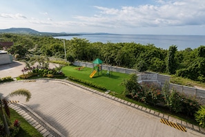 A playground with a slide and swings, surrounded by greenery and a view of the sea.