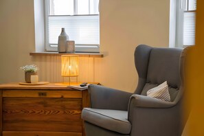 Ein Zimmer mit Schreibtisch aus Holz, einem grauen Sessel, einer Lampe, einer Vase und einem Fenster mit Jalousien.