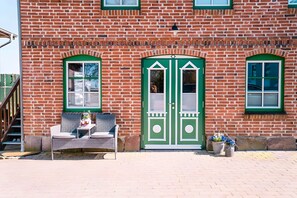 A brick building with a green door, two windows, a bench outside, and potted plants.