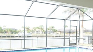 Área da piscina com estrutura de vidro, vista para um corpo d'água e espaço de estar com um banco acolchoado.