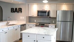 Uma cozinha moderna com armários brancos, uma ilha central, micro-ondas, frigorífico e luminária no teto.