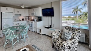 Una cocina moderna con armarios blancos, una mesa de comedor con sillas, un televisor de pantalla plana y vista al océano a través de amplios ventanales.