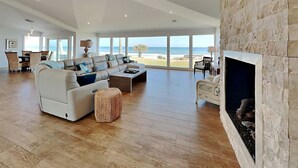 A spacious living room with a large window overlooking the beach, a fireplace, and a dining area with wicker furniture. - Ocean Sunrise | Luxury Group Retreat +  Atlantic Views (Ponte Vedra)