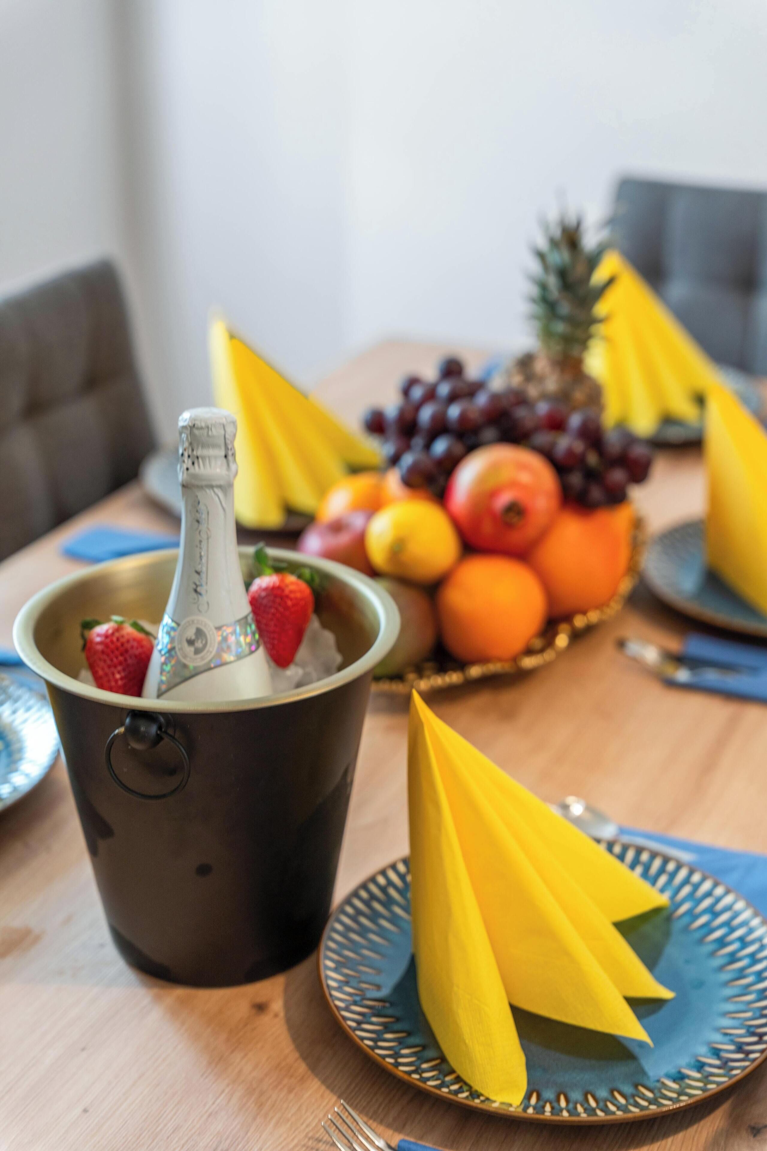 Een eettafel met een fles champagne in een emmer, een fruitschaal met verschillende soorten fruit, en gele servetten op borden.