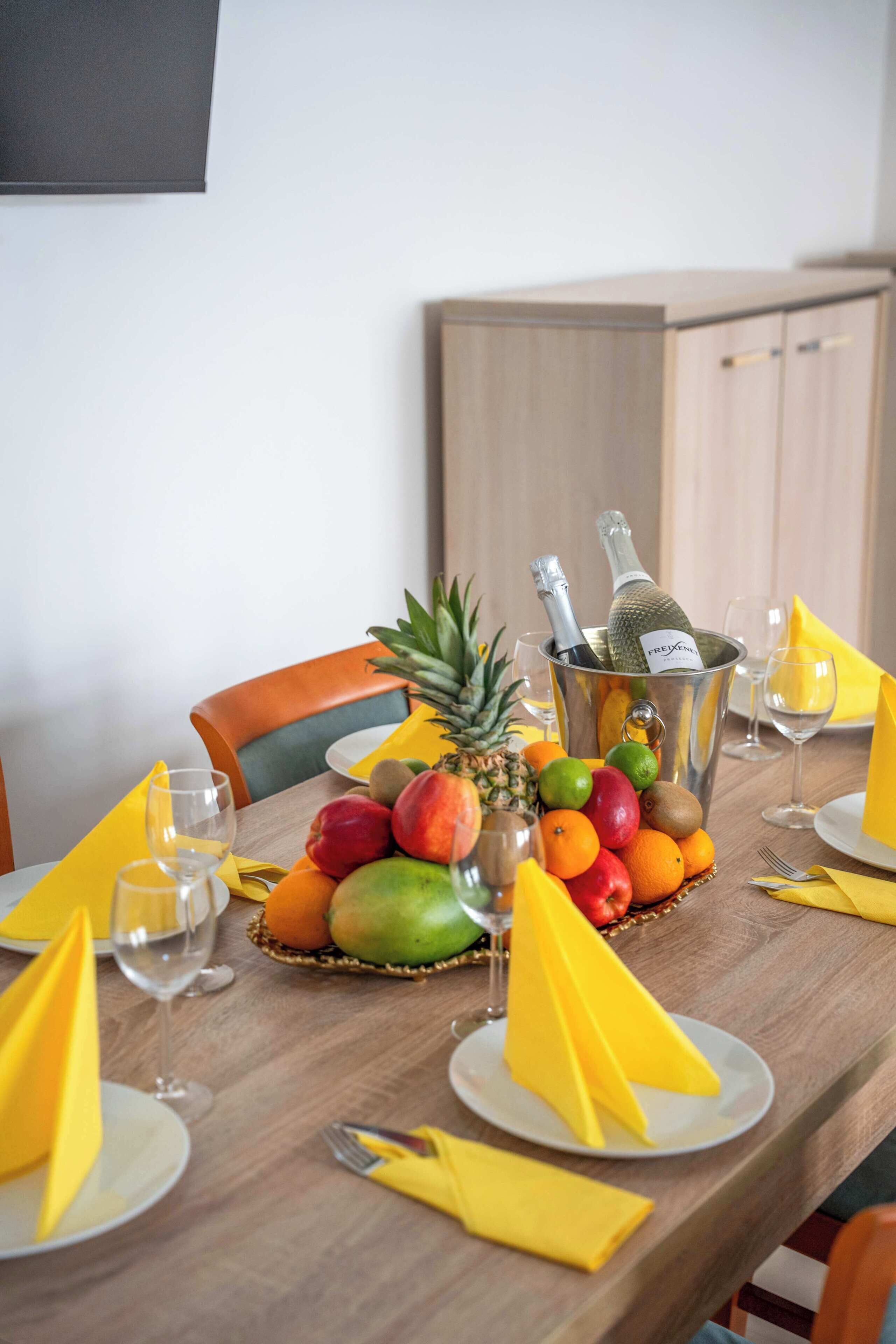A dining table set with fruit, a bottle of champagne, and yellow napkins.