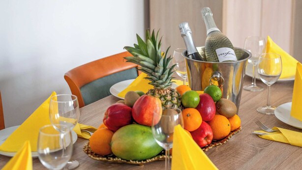 Ein Esstisch gedeckt mit Obst, einer Champagnerflasche und gelben Servietten.