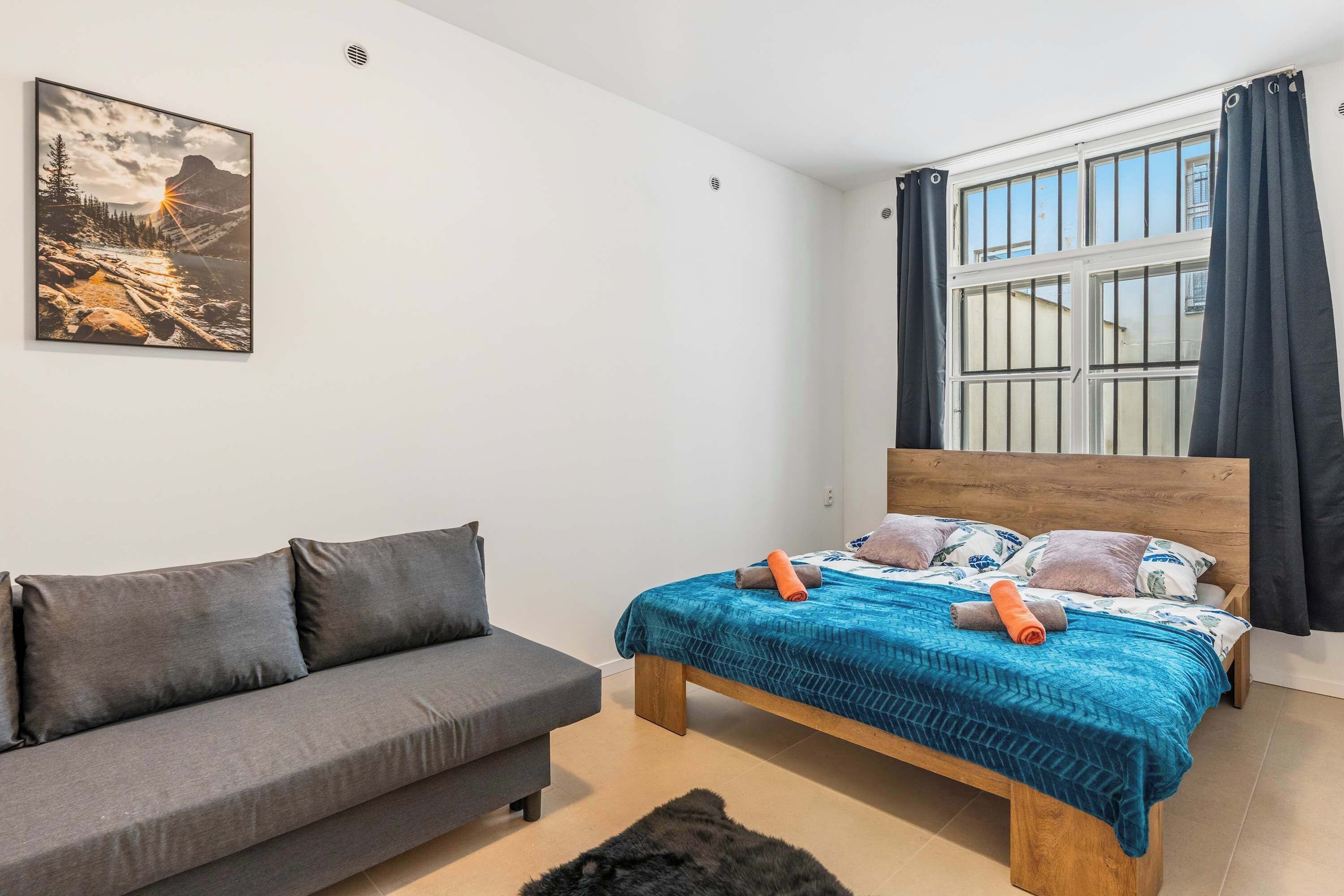 A bedroom with a bed, a grey sofa, a painting, and a window with curtains.