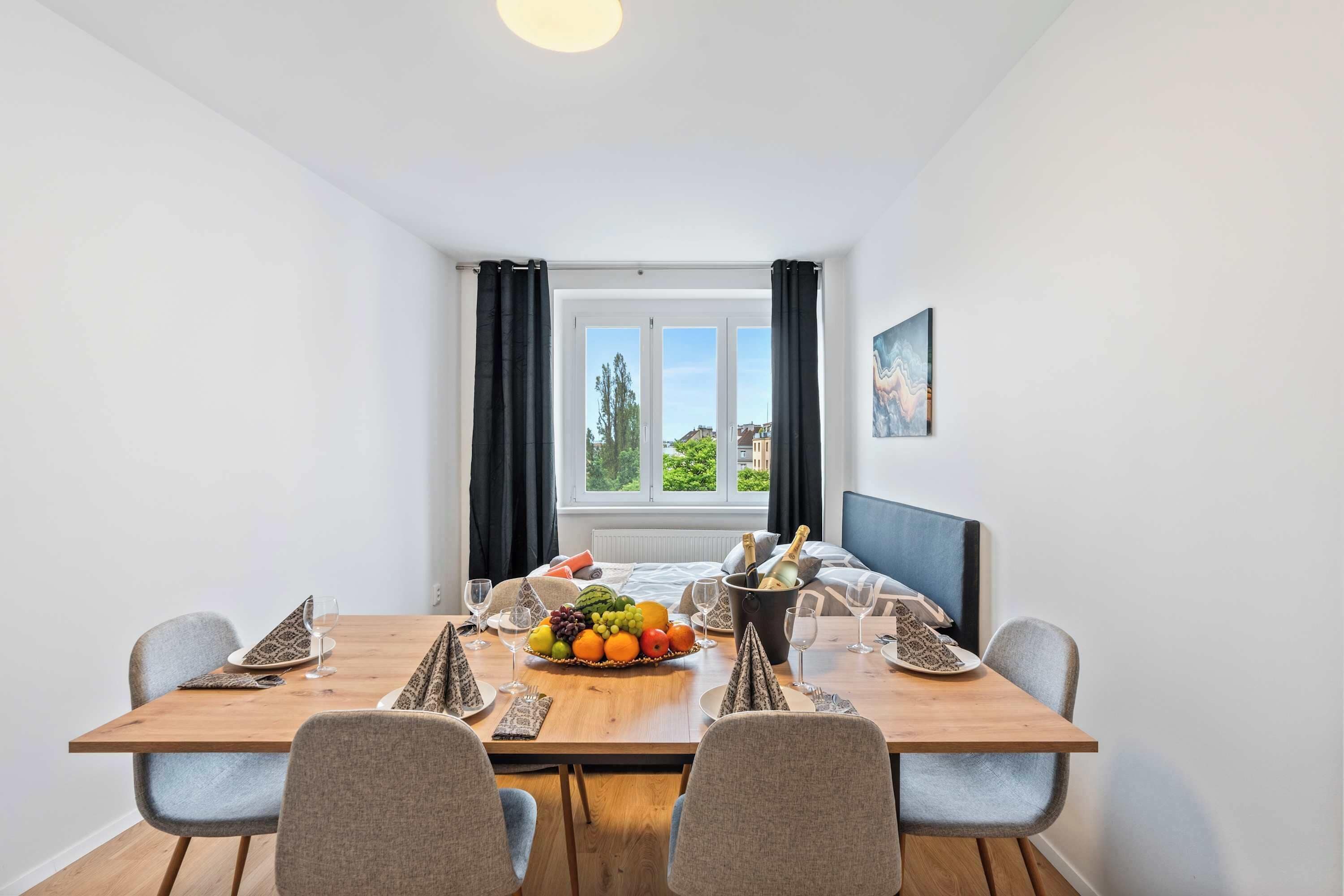 Un comedor con una mesa de madera, sillas grises, una cesta de frutas y una cama con cabecera azul al fondo.