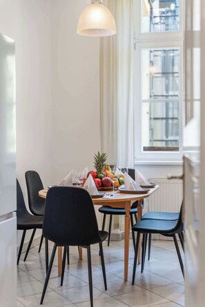 Uma área de refeições com uma mesa redonda, cadeiras pretas, uma cesta de frutas e um lustre.