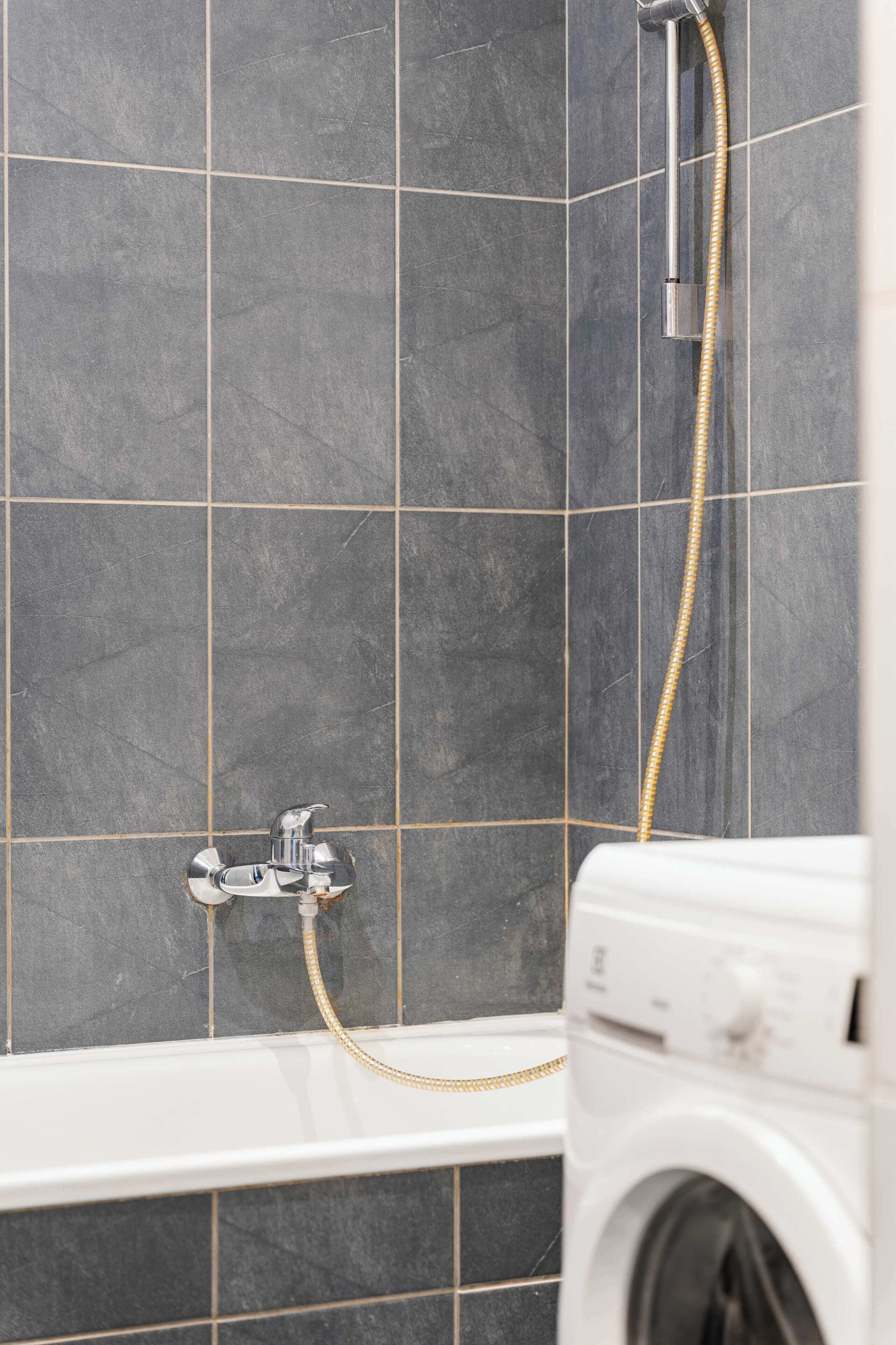 Un baño con baldosas oscuras, un cabezal de ducha, una manguera y una lavadora.