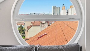 Un ventanal redondo con vistas a un paisaje urbano, un sofá gris con dos cojines, y un cielo azul brillante.