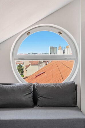 Een rond raam met uitzicht op een stadsbeeld, een grijze bank met twee kussens, en een helderblauwe lucht.