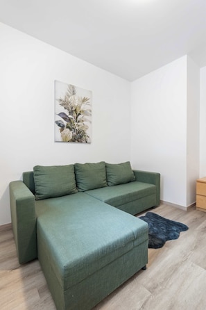 Ein grünes Ecksofa mit einem dunklen Teppich, Holzboden und einer Wandpflanze.