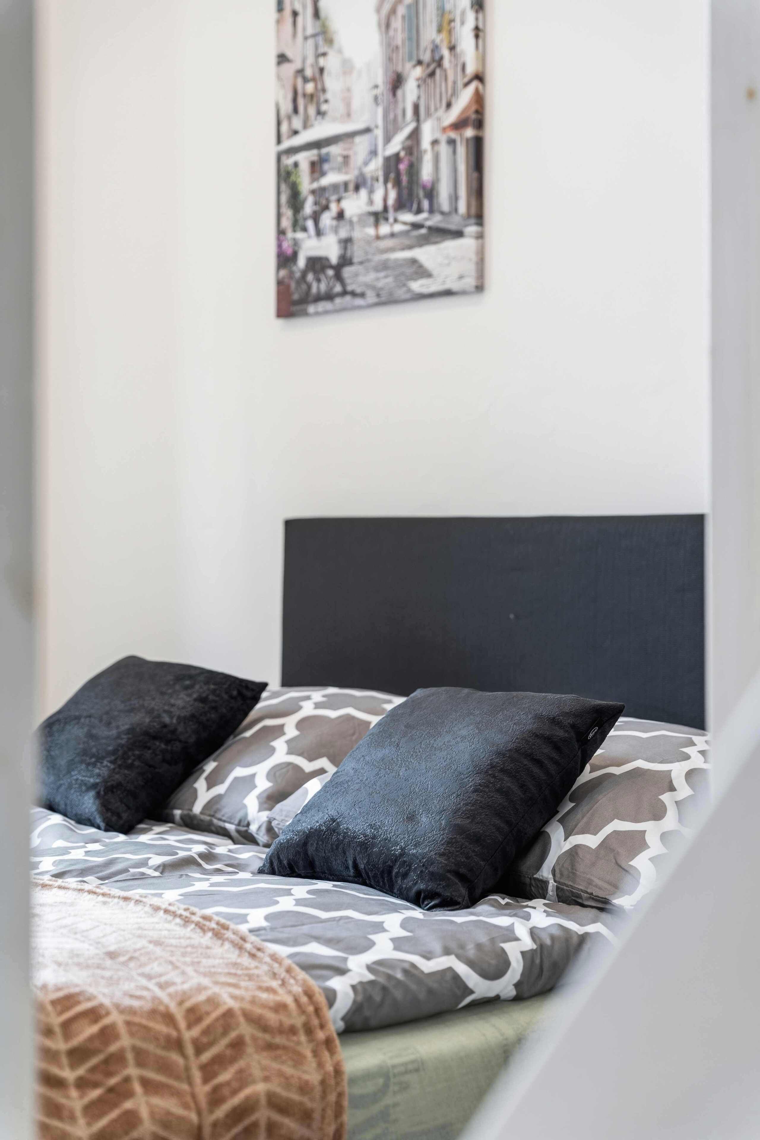Un lit avec une couvre-lit à motifs et deux oreillers, une tête de lit noire et un tableau représentant un paysage urbain accroché au mur.