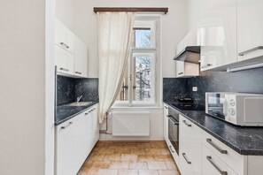 Une cuisine étroite avec des meubles blancs, un plan de travail noir, un four à micro-ondes et une fenêtre avec un rideau.