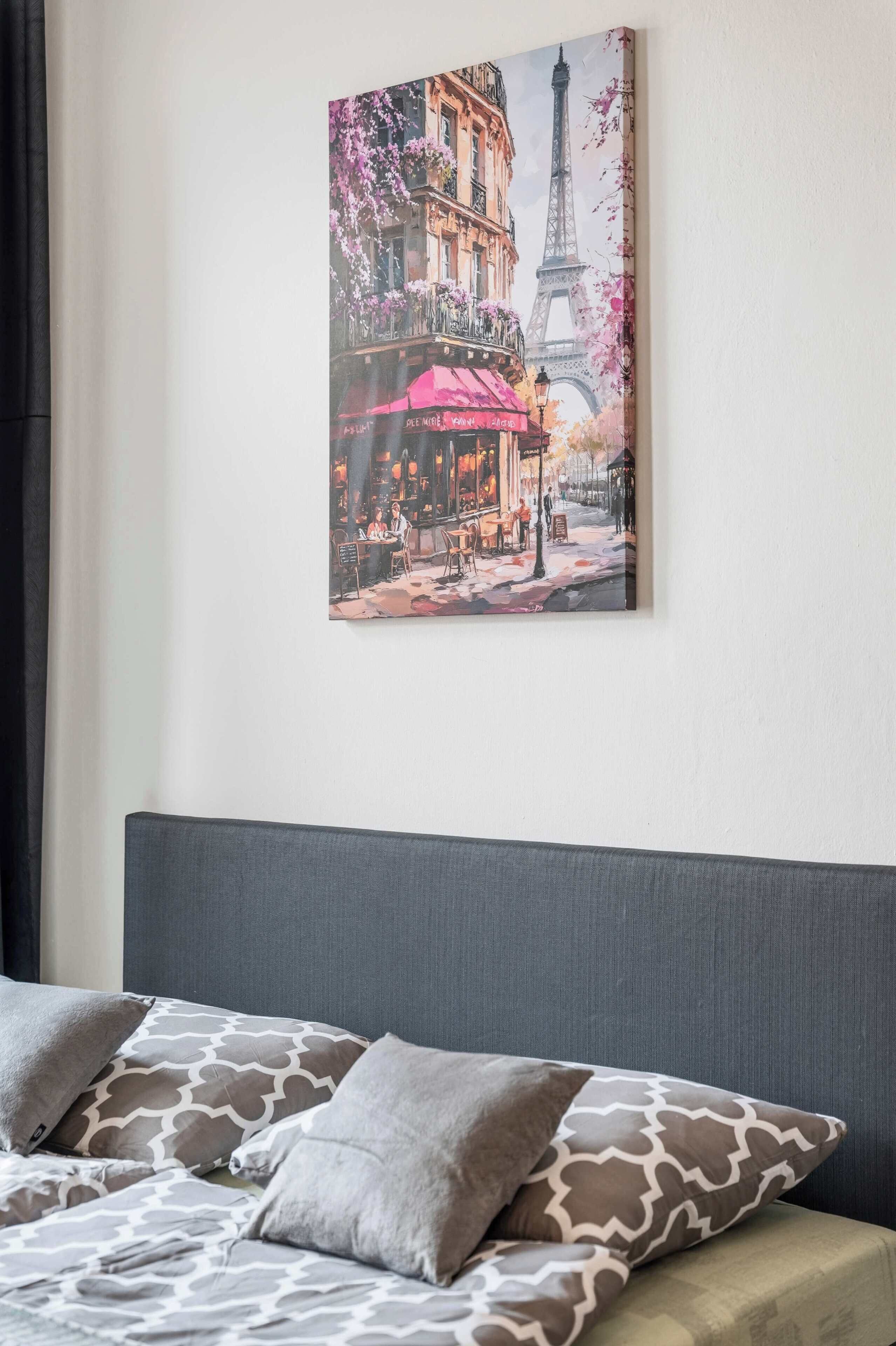 A bedroom with a bed, pillows, a headboard, and a wall-mounted painting of Parisian street scene.