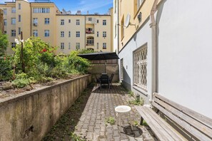 Ein schmaler Innenhof mit einem gepflasterten Weg, einem kleinen Tisch und Stühlen, einer Holzbank und einer Betonmauer.