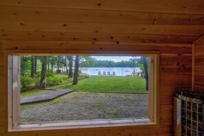 A wooden cabin with a window view of a grassy yard, a wooden deck, and a lake.